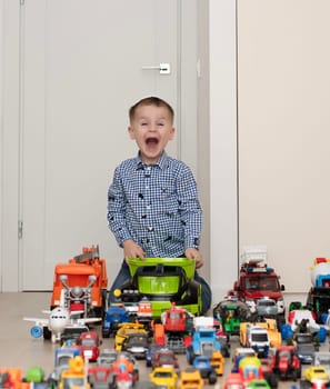 Concept of children's toys. A little boy, 4 years old, cheerful and handsome, happily plays, sitting on the floor, with multi-colored small and large cars in the children's room. Soft focus