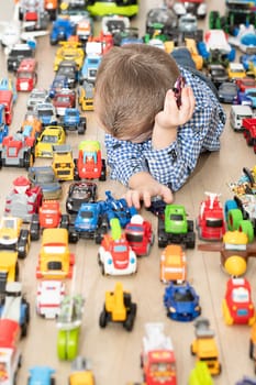 Concept of children's toys. A little boy, 4 years old, plays happily, lying on the floor, with colorful small and large cars in the children's room. Soft focus.