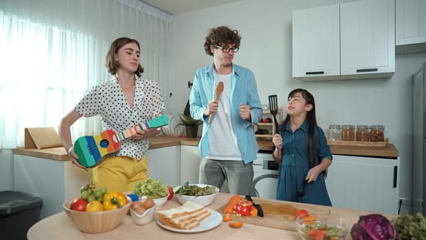 Caucasian skilled father, mother and asian daughter making breakfast while dancing together. Skilled mom playing ukulele while preparing vegetable at modern kitchen. Healthy food concept. Pedagogy.