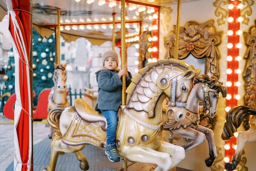 Little girl rides a toy horse on a carousel in the square near a decorated Christmas tree. High quality photo