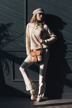 Studio portrait of beautiful woman with a curly blond hair holding black bag, posing on gray background. Model wearing stylish cap, sweater and classic trousers