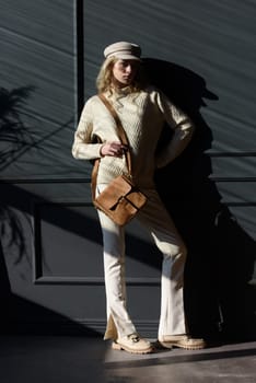 Studio portrait of beautiful woman with a curly blond hair holding black bag, posing on gray background. Model wearing stylish cap, sweater and classic trousers