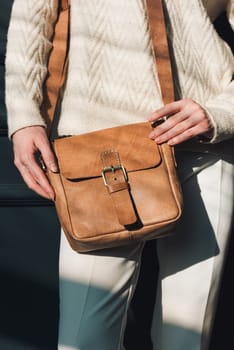 beautiful woman posing with a small brown bag near grey wall. Model wearing stylish white sweater and classic trousers