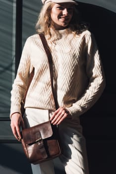 Studio portrait of beautiful woman with a curly blond hair holding brown bag, posing on gray background. Model wearing stylish cap, sweater and classic trousers