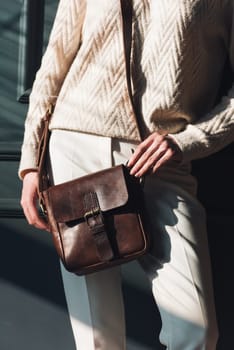 beautiful woman posing with a small brown bag near grey wall. Model wearing stylish white sweater and classic trousers