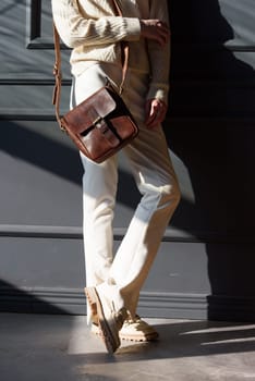 beautiful woman posing with a small brown bag near grey wall. Model wearing stylish white sweater and classic trousers