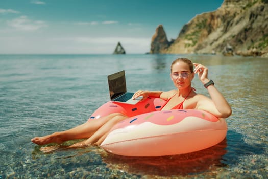 Woman laptop sea. Freelancer woman in sunglases floating on an inflatable big pink donut with a laptop in the sea. People summer vacation rest lifestyle concept