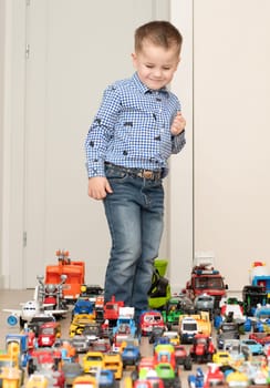 Children. Toy cars. A small cheerful and handsome boy, 4 years old, wearing a checkered shirt, plays with many colorful cars in a home interior. The toys are placed evenly on the floor.