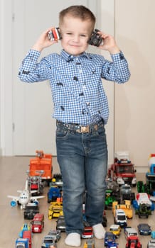 Children. Toy cars. A small cheerful and handsome boy, 4 years old, wearing a checkered shirt, plays with many colorful cars in a home interior. The toys are placed evenly on the floor.