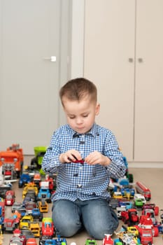 Concept of children's toys. A little boy, 4 years old, enthusiastically plays, sitting on the floor, with multi-colored small and large cars in the children's room. Soft focus