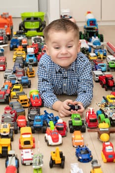 Concept of children's toys. A little boy, 4 years old, plays happily, lying on the floor, with colorful small and large cars in the children's room. Soft focus.