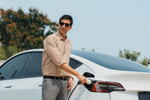 Young man recharge electric car's battery from charging station in outdoor green city park in springtime. Rechargeable EV car for sustainable environmental friendly urban travel lifestyle. Expedient