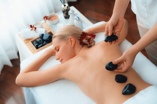 Hot stone massage at spa salon in luxury resort with day light serenity ambient, blissful woman customer enjoying spa basalt stone massage glide over body with soothing warmth. Quiescent