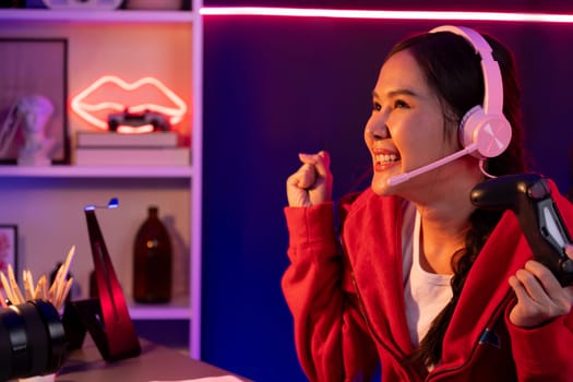 Host channel of smiling beautiful Asian girl streamer with joystick playing with raising fist up winning Esport skilled team players wearing headphones pastel media online in neon room. Stratagem.