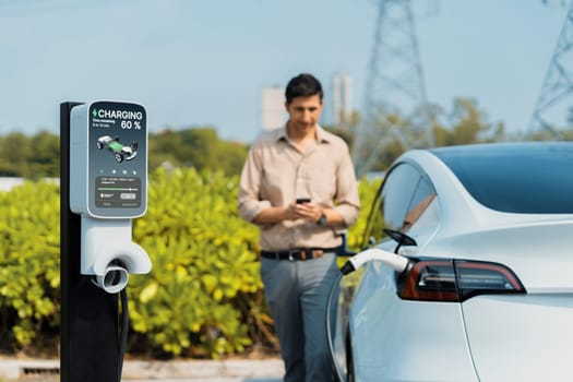 Man pay for electricity with smartphone while recharge EV car battery at charging station connected to power grid tower electrical as electrical industry for eco friendly car utilization.Expedient