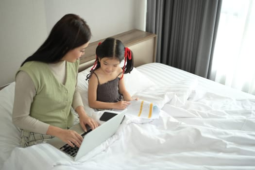 Beautiful mother working on laptop near her preschooler daughter in bedroom.