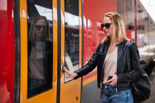 Young blond woman in jeans, shirt and leather jacket wearing bag and sunglass, presses door button of modern speed train to board on train station platform. Travel and transportation.