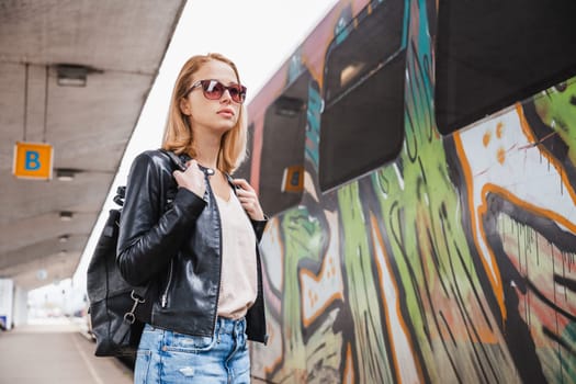 Young blond woman in jeans, shirt and leather jacket wearing bag and sunglass, embarking modern speed train on train station platform. Travel and transportation
