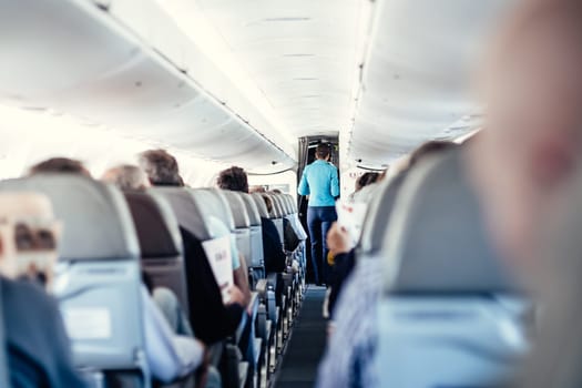 Interior of airplane with passengers on seats and stewardess in uniform walking the aisle, serving people. Commercial economy flight service concept