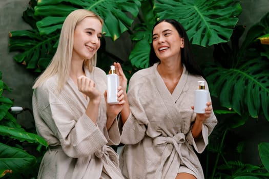 Tropical and exotic spa garden with bathtub in modern hotel or resort with two women in bathrobe holding beauty skincare product while enjoying leisure lush with greenery foliage background. Blithe