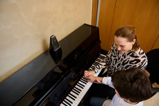 View from above of a Caucasian pleasant mature female music teacher, musician pianist teaching piano, explaining to n adolescent student boy the correct position of fingers on the piano keys