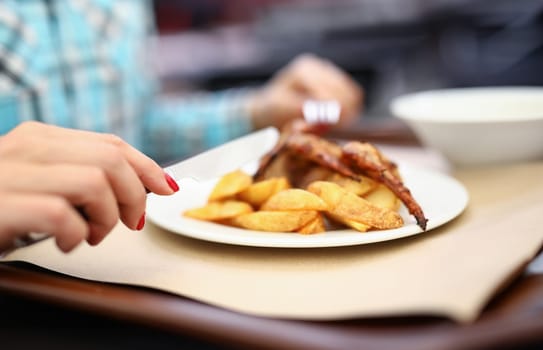 Woman hold knife and fork in her hand. White plate of potatoes and chicken on table.