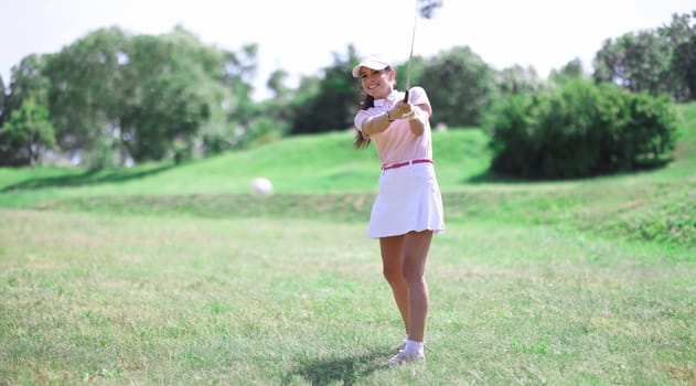 Beautiful woman in white skirt, cap and pink t-shirt play golf on green lawn. Close-up hitting ball with club.
