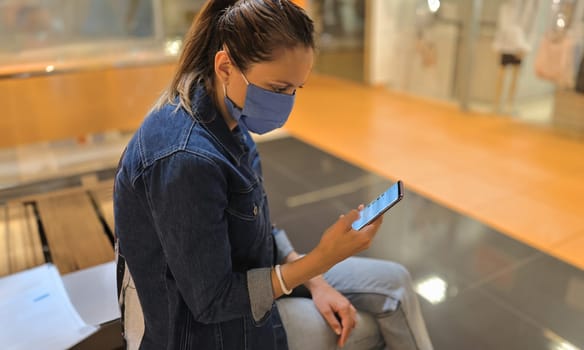 Woman in protective mask sit on bench in shopping center and look at phone. Empty hall in trading floor, showcases with manikin.