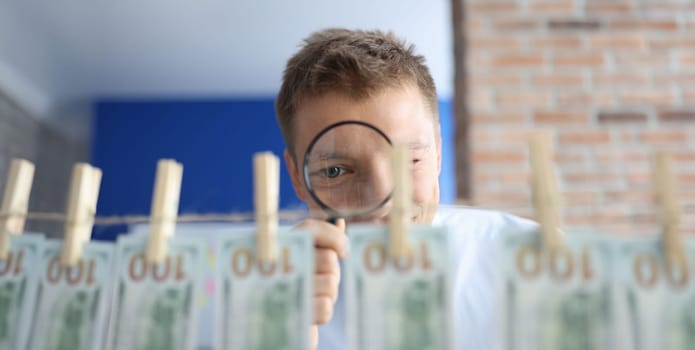 Man examines money through magnifying glass. Many hundred dollars hang on rope on clothespins.