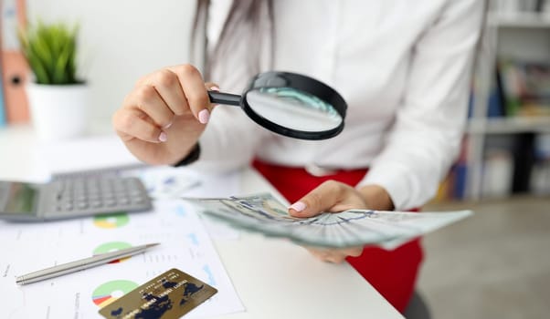 Woman check currency for authenticity. Female hand hold magnifying glass and lot of money. On white table lies gray calculator, metal pen, bank card and document with diagram.