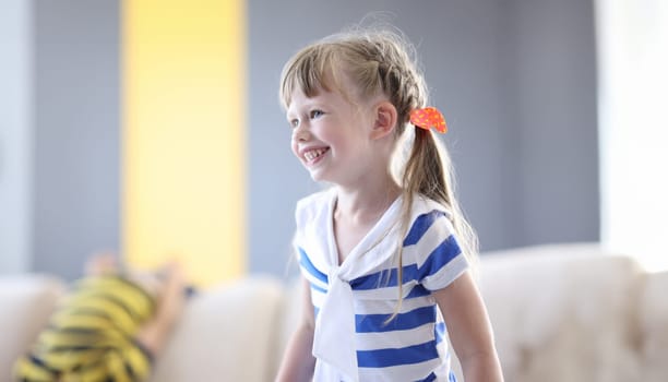 Happy girl in striped T-shirt and with pigtails laugh. In background child indulge on couch.