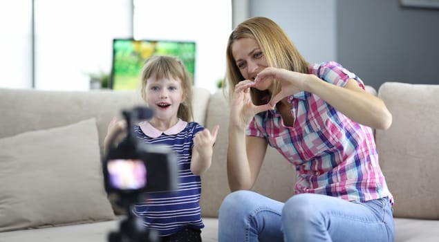 Woman with child is sitting on couch in apartment and showing hand gesture in front of camera. Family business blogging concept.