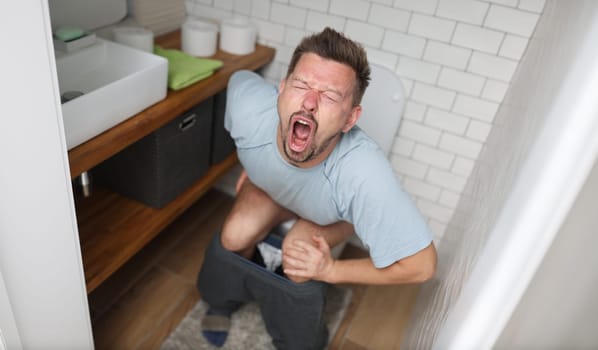 Young man with suffering on his face sits on toilet in bathroom. Indigestion constipation concept.