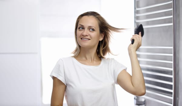 Young beautiful woman is looking at her reflection in bathroom and combing her hair. Woman hair styling at home concept.