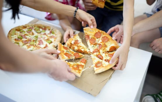 Delicious pizza close-up cut into pieces on white table. Family with children eat pizza and take pieces with their hands.