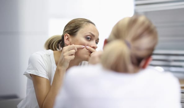 Young woman stands in front of mirror and presses pimples with hands portrait. Facial skin care cleaning at home concept.