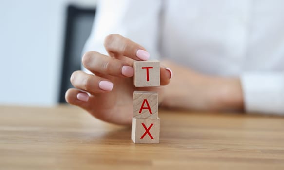 Female hand build tower from wooden cubes on table. Woman composes word tax from letters.