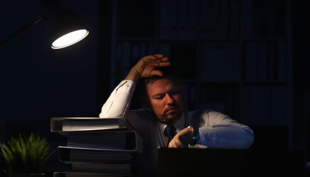 Portrait of exhausted businessman sitting indoors and looking at expensive watches trying to skip overtime job and holding hand near forehead. Night at office concept