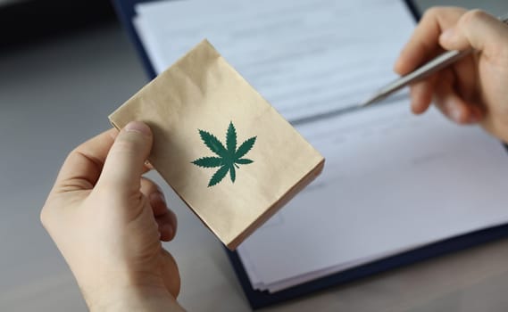 Close-up of person holding package with cannabis. Patients hand signing paper at doctors office. Meds for therapy. Traditional medicine and healthcare concept
