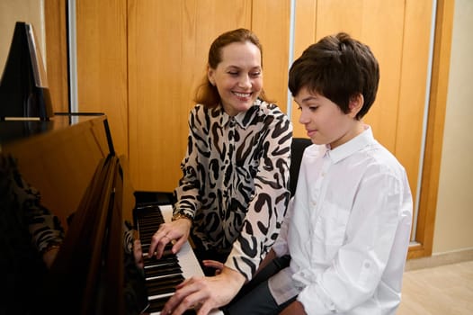 Smiling woman playing piano, sitting near his adolescent son, performing Christmas song, enjoying the rhythm of melody. Happy family of a cute teenage boy and his mother playing grand piano together