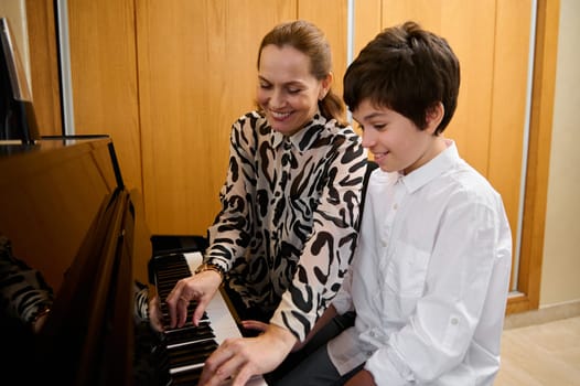Authentic portrait of a female pianist musician holding the hands of a teenage boy, showing the true position of finger on piano keys, explaining the piano lesson during individual music class indoors