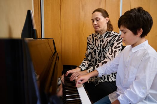 Authentic portrait of a Caucasian young adult loving mother and son sitting at grand piano, performing classic melody, toucher white and black keyboard of the piano forte, playing piano, singing song