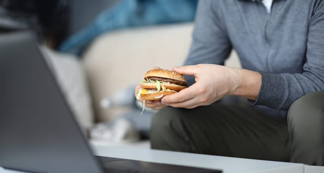 Focus on male hand holding unhealthy sandwich and preparing to consume fat dish full of quick carbohydrates and mischievous calories. Harmful fastfood concept