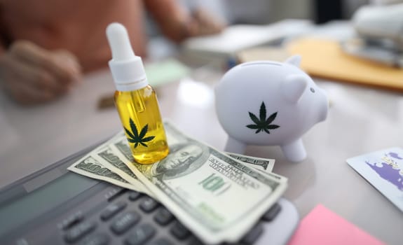 Close-up of white piggy bank with marijuana sign, cannabinoid oil in bottle with pipette. Cash on table. Person paying for pain medication. Painkillers concept