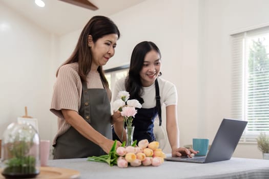 Senior mother and adult daughter happy on the table while arrange flowers in a vase together using laptop computer. lifestyle concept. Happy time together.