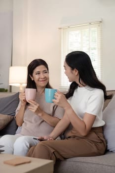 Happy senior mother with adult daughter sitting on couch and holding cups with coffee or tea at home. Enjoy family concept.