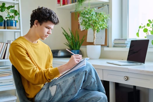 Guy college student sitting at home at desk looking at laptop screen, making notes in notebook. Young male studying online, watching webinar, browsing Internet. E-learning, education, technology youth