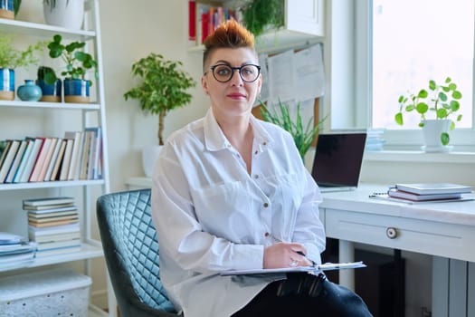 Portrait of smiling female psychotherapist with clipboard at workplace in office. Professional mental therapist counselor psychologist social worker looking at camera. Health care services, treatment