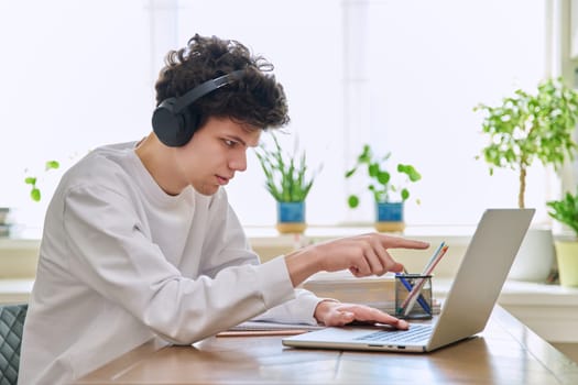 Young male student in headphones sitting at home at desk looking talking in web camera of computer laptop. Guy 19-20 years old studying remotely, video conference call, e-learning technology education