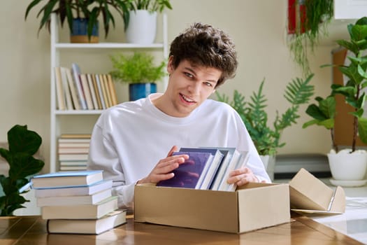 Satisfied young male customer buyer sitting at home unpacking cardboard box parcel with new books, online purchases. Delivery by mail, internet store bookstore